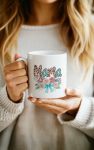 A white ceramic mug with a stylish "Mama" design in leopard print, accented with pearls, roses, and a teal bow.